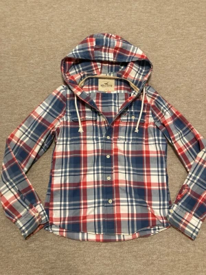 Rutig hoodie från Hollister - Snygg röd, blå och vit rutig hoodie från Hollister med knappar framtill och dragsko i huvan. Långärmad modell i mjukt bomullstyg, perfekt för en avslappnad och trendig look. Klassisk ficka på bröstet och bekväm passform.