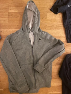 Grå Nike Therma-Fit hoodie XL - Säljer en grå zip-hoodie från Nike i modellen Therma-Fit. Hoodien har dragkedja framtill, två fickor och en mjuk, fleecefodrad insida. Klassisk Nike-logga broderad på bröstet och stor huva. Perfekt för chill eller träning.