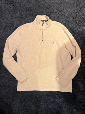 Polo Ralph Lauren halvzip - Snygg beige långärmad tröja från Polo Ralph Lauren med halvzip och liten broderad logga på bröstet. Tröjan har hög krage och är tillverkad i mjukt material, perfekt för lager på lager. Enkel och stilren design som passar till mycket.