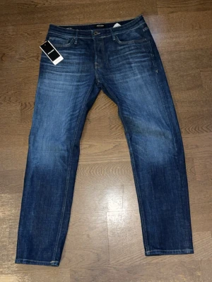 Helt nya mörkblåa tapered jeans från Jack & Jones - Säljer ett par fina mörkblå tapered jeans från Jack & Jones med klassisk femficksdesign och snygga slitningar. Jeansen har normal midja, raka ben och är tillverkade i stretchigt denim för extra komfort. Perfekta för en avslappnad och trendig look. Modellen är tapered.