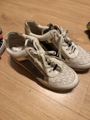 Vita och guldiga sneakers Michael Kors - Snygga sneakers från Michael Kors i vitt och guld med klassiskt MK-mönster. Skorna har vita snören, gulddetaljer och paneler i beige och brunt tyg. Perfekta för dig som vill ha en trendig och lyxig look till vardags. Tydlig MK-logga på hälen.