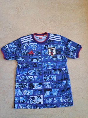 Blå Japan Adidas fotbollströja - Unik blå fotbollströja från Adidas och Japan med manga-inspirerat tryck i olika blå nyanser. Tröjan har korta ärmar, rund hals med röd kant och Japans fotbollsförbunds logga på bröstet. Perfekt för dig som älskar både fotboll och anime.