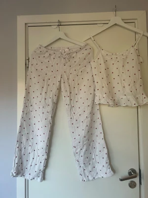 Vitt pyjamas-set med hjärtmönster💕💕 - Säljer ett pyjamas-set i vitt med små röda hjärtan💕💕💕Setet består av ett linne med tunna axelband och volangkant samt ett par lösa byxor med resår i midjan💕💕💕. Materialet är mjukt och luftigt, perfekt för att chilla hemma med💕💕💕💕I bra skick💕💕Jag säljer  båda byxorna och linnet för 100 kr tillsammans💕💕💕Hör av er vid intresse💕💕💕💕