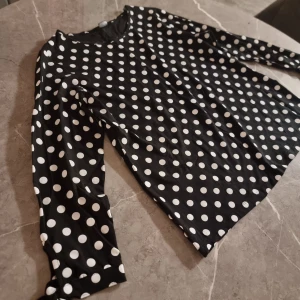 Svart prickig långärmad topp - Snygg svart långärmad topp med vita prickar och rund hals. Tillverkad i ett mjukt och stretchigt material som sitter bekvämt. Perfekt för dig som gillar klassiska mönster med en lekfull twist.