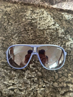 Blå Carrera pilotbågar solglasögon - Snygga solglasögon från Carrera med blå pilotbågar och mörka tonade glas. Klassisk aviatorform med en cool detalj i metall vid näsryggen. Perfekta för dig som vill ha en sportig och trendig look i sommar.