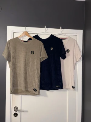 T-shirt trio från ACE i olika färger - Säljer tre stilrena t-shirts från ACE i beige, mörkblå och ljusgrå. Alla har rund hals, korta ärmar och ACE-logga på bröstet samt en liten patch vid nederkanten. Materialet ser ut att vara mjukt och bekvämt, perfekt för en avslappnad look.