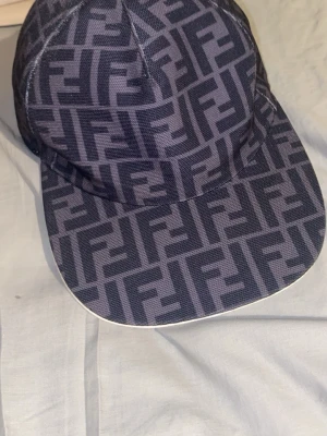 Fendi keps med FF-mönster - Säljer en snygg keps från Fendi med det klassiska FF-logomönstret i svart och grått. Kepsen har böjd skärm och är tillverkad i ett tåligt tygmaterial. Perfekt för dig som vill ha en trendig och exklusiv accessoar.