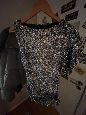 Glittrig silver kortklänning - Säljer en glittrig kortklänning i silver med massor av paljetter. Klänningen har en asymmetrisk design med en ärm och en axel bar. Perfekt för dig som vill sticka ut och glänsa på nästa fest eller event!