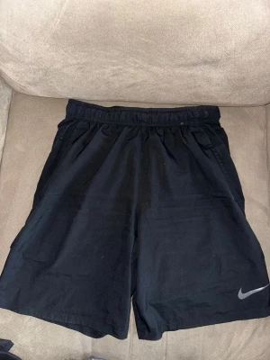 Svarta Dri-FIT shorts från Nike - Säljer ett par svarta Nike Dri-FIT shorts med elastisk midja och diskret logga på benet. Perfekta för träning eller chill dagar. Tillverkade i ett lätt och snabbtorkande syntetmaterial som håller dig bekväm hela dagen.