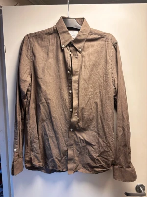 Beige långärmad skjorta med knappar - Stilren beige skjorta med klassisk krage och knappar framtill. Skjortan har långa ärmar och är tillverkad i ett lätt bomullstyg som känns skönt mot huden. Perfekt för dig som gillar en enkel och clean look.