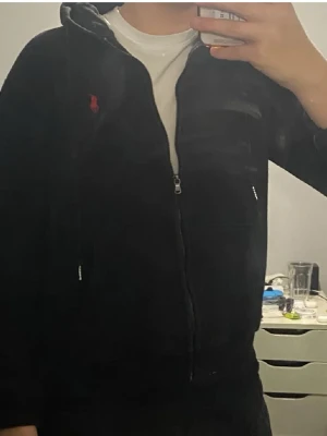 Svart hoodie från Polo Ralph Lauren - Säljer en svart hoodie med dragkedja från Polo Ralph Lauren. Klassisk design med huva, dragsnören och kängurufickor. Liten röd logga broderad på bröstet. Tillverkad i mjukt bomullsmaterial som är skönt att ha på sig.