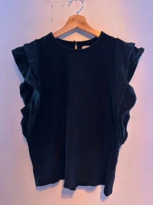 Svart topp med volangärmar - Snygg svart topp med breda volangdetaljer längs ärmhålen. Toppen har rund halsringning och är ärmlös, vilket ger en modern och trendig look. Perfekt att styla med jeans eller kjol för en cool outfit.