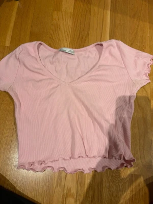Ljusrosa ribbad croppad topp - Söt ljusrosa croppad topp från pull &bear med ribbad struktur och v-ringning. Toppen har korta ärmar och vågig volangkant nedtill och vid ärmsluten. 