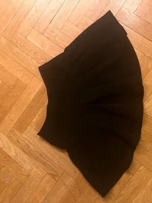 Svart kortkjol med volang - Säljer en svart kortkjol med volangkant. Kjolen har en utsvängd form som ger en snygg siluett och passar perfekt till en trendig outfit. Materialet känns mjukt och följsamt, troligen polyester. Enkel att matcha med olika toppar och skor.Skriv gärna för mått! 💕