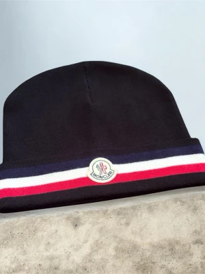 Svart Moncler mössa med ränder - Snygg svart mössa från Moncler med vikbar kant och klassiska Moncler-loggan framtill. Mössan har en randig detalj i marinblått, vitt och rött runt kanten. Tillverkad i mjukt stickat material som håller dig varm och stilren.