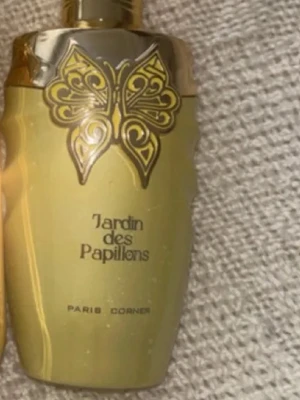 Jardin des Papillons parfymflaska - Elegant parfymflaska i guld med dekorativ fjäril på framsidan och texten 'Jardin des Papillons'. Flaskan har en glänsande finish och exklusiv design som sticker ut i badrumshyllan.