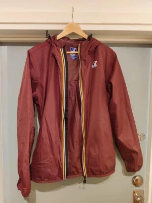 Vinröd vindjacka från K-Way M - Säljer en vinröd vindjacka från K-Way med huva och dragkedja med gul och vit kant. Jackan är tillverkad i lätt och vattenavvisande material, perfekt för blåsiga dagar. Klassisk logotyp på bröstet och justerbar huva. Långärmad och enkel design.