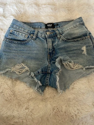 Blåa slitna jeansshorts från 157 - Säljer ett par blå denimshorts från 157 med hög midja och råa, fransiga kanter. Shortsen har slitningar framtill och klassisk femficksdesign. Perfekta för sommaren och ger en avslappnad vibe. Stolen xxs , pris 20kr 