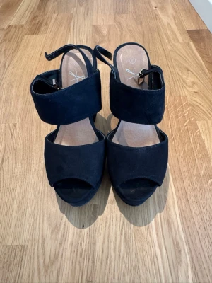 Svarta chunky pumps med bred klack - Säljer ett par svarta pumps med bred klack och platåsula. Skorna har två breda band över foten och öppen tå, samt justerbar rem runt ankeln. Materialet är mockaimitation som ger en snygg och trendig look. Perfekta för dig som gillar statement-heels.