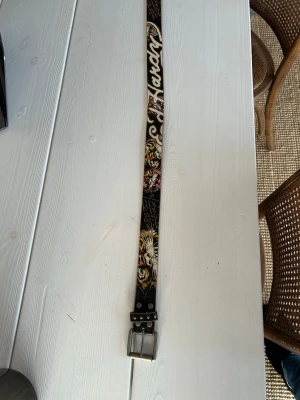 Bälte från Ed Hardy - Coolt svart bälte med färgglada tigertryck och vit text längs hela bandet. Bältet har en silverfärgad metallspänne och detaljer i nitar. Perfekt för att ge din outfit en edgy vibe. Bältet är 113 cm