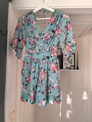 Blommig omlottshortsklänning från TFNC - Supersöt ljusblå omlottshortsklänning från TFNC London med blommigt mönster i rosa, vitt och grönt. Klänningen har v-ringning, knyt i nacken och korta ärmar. Perfekt för sommarens alla tillfällen!