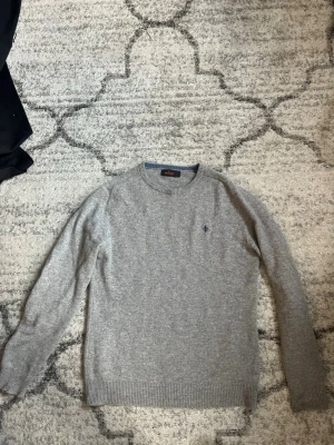 Grå stickad tröja från Morris - Klassisk grå stickad tröja från Morris med rund halsringning och diskret blå logga broderad på bröstet. Tröjan har ribbade muddar vid ärmslut och nederkant. Perfekt basplagg för lager på lager och enkel att matcha.