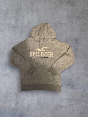 Hollister hoodie  - Snygg gråmelerad hoodie från Hollister med stor logga och texten 'California' på bröstet. Klassisk känguruficka framtill och justerbar huva med dragsko. Perfekt för en avslappnad och trendig look.