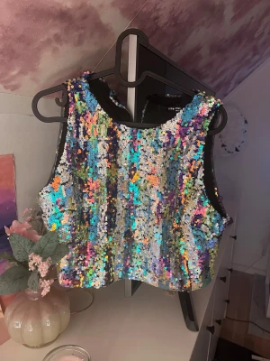 Regnbågsglitter crop top med paljetter - Säljer en färgsprakande crop top täckt av skimrande paljetter i regnbågens alla färger. Toppen har breda axelband och rund halsringning, perfekt för dig som vill sticka ut. Materialet är tätt med paljetter på svart tygbas för extra effekt. Absolut inga fel på den och har haft på mig den 2 ggr.
