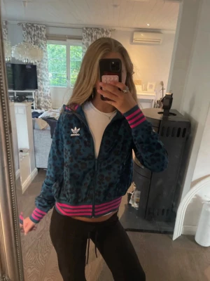 Adidas leopardmönstrad ziptröja - Snygg ziptröja från Adidas med blått och svart leopardmönster. Tröjan har dragkedja, ribbade muddar och midja i rosa och svart randigt. Adidas-logga broderad på bröstet. Som ny