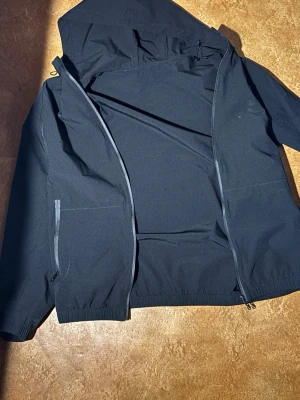 Svart vindjacka med huva - Stilren svart vindjacka med huva och hel dragkedja framtill. Jackan har två sidofickor med dragkedja och elastiska muddar vid ärmsluten. Materialet är tunt och smidigt, perfekt för blåsiga dagar. Enkel design utan synligt märke.