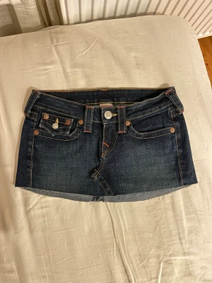 Mörkblå jeanskjol från True Religion - Säljer en mörkblå kort jeanskjol från True Religion med klassiska kontrastsömmar och dekorativa fickor både fram och bak. Kjolen har bälteshällor, knapp- och dragkedjestängning samt rå kant nertill för en unik look.passar s/xs