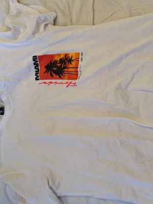 Vit t-shirt med Miami-tryck - Säljer en vit t-shirt med färgglatt Miami-tryck på bröstet. Trycket visar palmer mot en solnedgång i orange och rött, samt texten 'Miami' och 'Florida'. Klassisk rund hals och korta ärmar. Perfekt för dig som gillar streetstyle och somriga vibes.