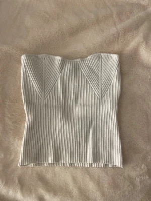 Vit ribbad tubtopp - Säljer en snygg vit ribbad tubtopp med markerade sömmar framtill som ger en cool struktur. Toppen är axelbandslös och har en tight passform som framhäver figuren. Perfekt att styla med jeans eller kjol för en trendig look.