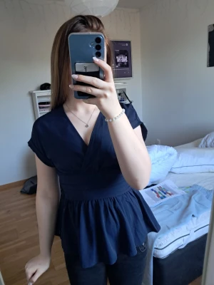 Mörkblå omlottopp med v-ringning - Snygg mörkblå omlottopp med v-ringning och korta ärmar. Toppen har markerad midja och ett lätt utställd nederdel som ger en feminin siluett. Perfekt att styla med jeans eller kjol för en trendig look.