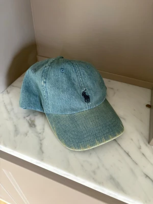 Ljusblå denimkeps från Polo Ralph Lauren - Säljer en ljusblå keps i denim från Polo Ralph Lauren med klassisk broderad logga framtill och 'POLO' broderat baktill. Justerbart spänne bak för perfekt passform. Snygg och avslappnad stil som passar till det mesta.