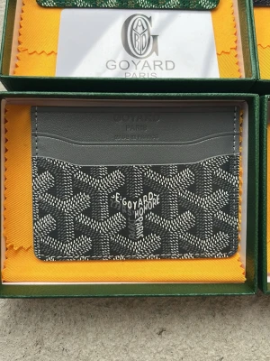 Grå plånbok från Goy4ard - Snygg grå korthållare från Goy4ard med klassiskt mönster i svart och vitt. Tillverkad i läder med flera fack för kort. Perfekt accessoar för dig som vill ha något stilrent och exklusivt i fickan.