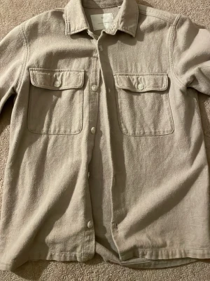 Beige overshirt med fickor - Stilren beige overshirt med två stora bröstfickor med lock och knappar. Skjortan har klassisk krage och knäppning framtill. Materialet ser ut att vara mjukt och borstat, perfekt för lager på lager. Enkel och clean design som passar till det mesta.