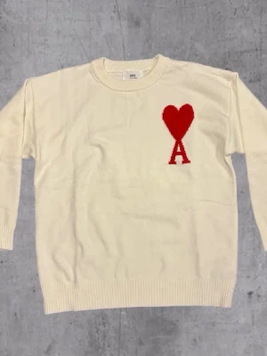 Vit stickad tröja från Ami Paris - Vit stickad tröja från Ami Paris med röd hjärtlogga och bokstaven A på bröstet. Tröjan har rund halsringning, långa ärmar och ribbade muddar vid ärmslut och nederkant. Perfekt för dig som gillar stilrena plagg med en twist.