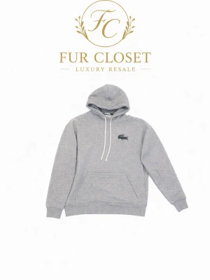 Grå hoodie från Lacoste med huva - Klassisk grå hoodie från Lacoste med huva och vita dragsnören. Ikoniska gröna krokodilloggan på bröstet och en ficka framtill. Tillverkad i mjukt sweatshirtmaterial som är perfekt för chill dagar. Ribbade muddar vid ärmslut och nederkant.