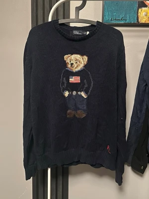Mörkblå stickad tröja Polo bear Ralph lauren  - Mörkblå stickad tröja från Polo Ralph Lauren med ikonisk Polo Bear på bröstet, björnen bär en tröja med amerikanska flaggan. Rund halsringning och ribbade muddar. RL-broderi vid nederkanten. Perfekt för dig som gillar klassisk preppy stil. Obs rep och är en riktig M passform lite oversized så passar även L, billigare vid snabb affär