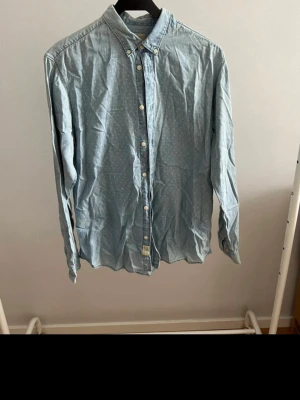 Ljusblå prickig skjorta med knappar - Snygg ljusblå skjorta med diskreta vita prickar och klassisk krage. Skjortan har långa ärmar och knäppning framtill. Tillverkad i mjuk bomull som känns skön mot huden. Perfekt för en avslappnad men ändå stilren look.