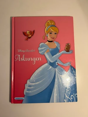 Askungen Disney klassiker bok - En färgglad och klassisk barnbok med hårda pärmar, rosa omslag och illustration av Askungen i blå klänning. Boken är en del av Disney klassiker-serien och berättar sagan om Askungen. Perfekt för dig som gillar magiska berättelser och Disney.