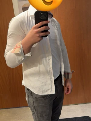 Vit skjorta från Burberry Brit - Klassisk vit skjorta från Burberry Brit med ikoniskt rutigt mönster på insidan av kragen och manschetterna. Skjortan har knappar framtill, lång ärm och är tillverkad i mjuk bomull. Perfekt för en clean och stilren look.