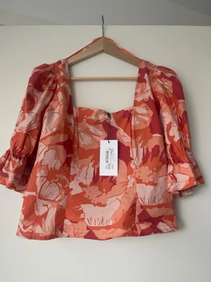 Blommig topp från Adoore i orange - Trendig topp från Adoore med blommigt mönster i orange, rött och beige. Modellen har puffiga trekvartsärmar med volangdetalj och fyrkantig ringning. Perfekt för dig som vill sticka ut med färg och mönster. Tillverkad i bomull för en skön känsla.