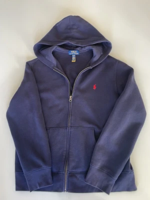 Mörkblå hoodie från Polo Ralph Lauren - Mörkblå hoodie med dragkedja från Polo Ralph Lauren. Klassisk design med huva, kängurufickor och det ikoniska röda Polo-logotypen broderad på bröstet. Tillverkad i mjukt bomullsmaterial som känns skönt mot huden. Perfekt för en avslappnad och stilren look.