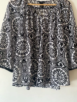 Svartvit mönstrad blus från H&M - Säljer en luftig svartvit blus från H&M med grafiskt cirkelmönster. Blusen har trekvartsärm och en lös, flowy passform. Materialet är tunt och genomskinligt, perfekt för lager på lager. Rund halsringning med liten rynkdetalj framtill.
