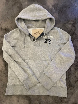 Hollister Hoodie - Snygg grå hoodie från Hollister med trycket 'Hollister 22' på bröstet. Hoodien har huva med dragsko, knappdetalj vid halsen. Tillverkad i mjukt bomullsmaterial som är skönt mot huden. Perfekt för en avslappnad och trendig look.