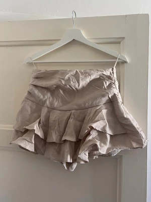 Beige volangskort med omlottkänsla - Supersnyggt beige skort med volanger och omlottkänsla i strl M. Skortsen har hög midja och är tillverkad i ett lätt, mjukt tyg som ger en flowy look. Perfekt för dig som vill ha kjolkänsla men ändå shortsens bekvämlighet. Endast testad.