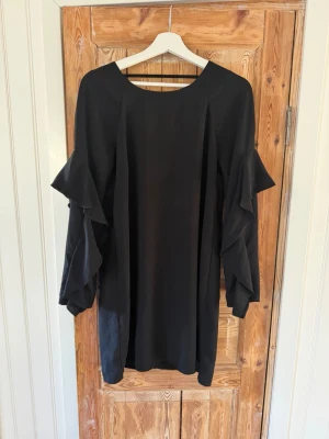 Svart klänning med volangärmar - Svart kortklänning med långa volangärmar och v-ringad rygg. Klänningen har en loose passform och är tillverkad i ett mjukt, lätt material som ger ett snyggt fall. 