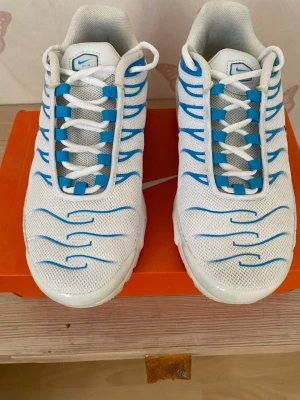 Nike Air Max Plus vita/blå skor - Endast använda ett fåtal gånger. storlek 41. Nike Air Max Plus sneakers i vitt mesh med blå detaljer och svarta swoosh-loggor. Skorna har synliga Air-enheter i sulan och blå vågformade linjer på ovandelen. Klassisk siluett med vita snören och bekväm dämpning. Perfekta för dig som gillar streetwear och sportig stil.